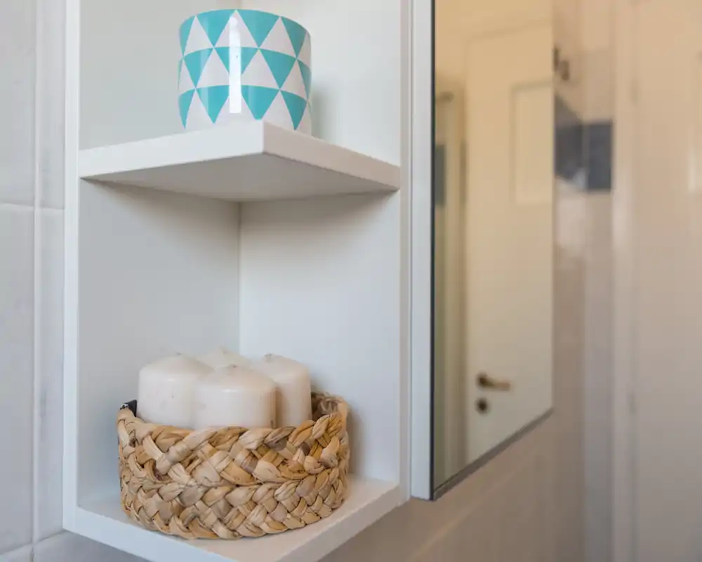 Dettaglio di una mensola del bagno con candele decorative in un cestino intrecciato e una tazza decorata; si vede parzialmente un pensile con specchio e la porta del bagno sullo sfondo.