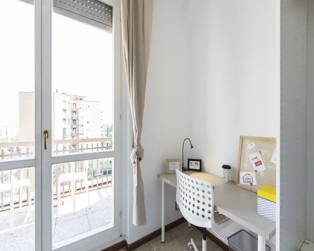 Pequeño espacio de trabajo luminoso junto al balcón: escritorio minimalista con silla, lámpara y corcho decorativo junto a grandes puertas de vidrio que dan a un balcón con vistas a la ciudad.