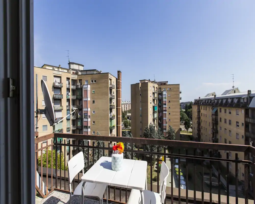 Balcón soleado con una pequeña mesa blanca y sillas, perfecto para el café de la mañana con vistas a los edificios vecinos.