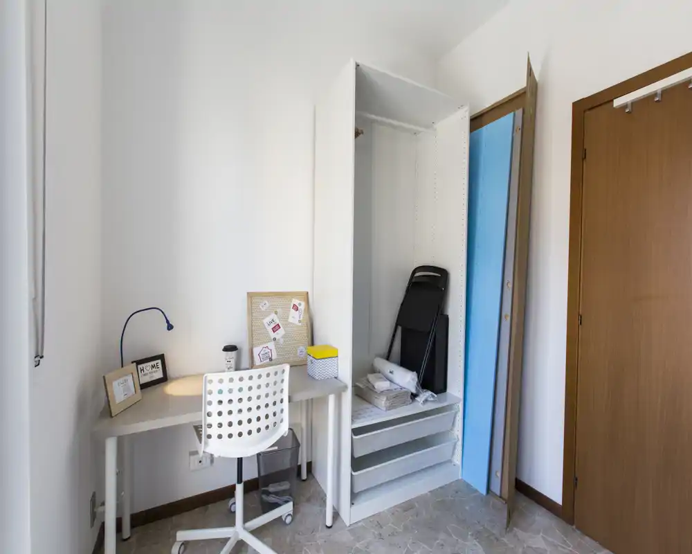 Pequeña habitación luminosa preparada como espacio de trabajo: escritorio compacto con silla, lámpara de trabajo y corcho junto a un armario abierto. Estilo limpio y luz natural, adecuado para estudiar o teletrabajar.