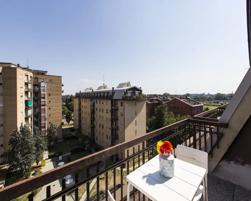 Balcón soleado con una pequeña mesa blanca y dos sillas, jarrón decorativo con flores y amplia vista a los edificios residenciales — ideal para tomar café por la mañana.