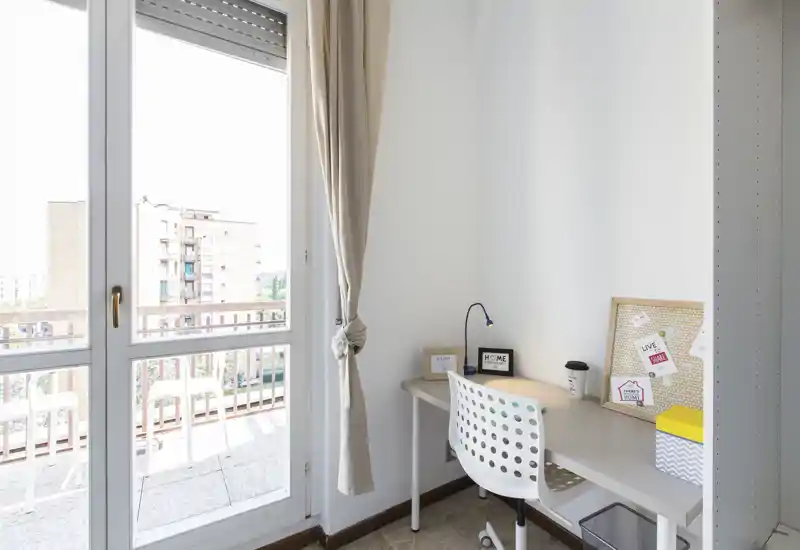 Pequeño espacio de trabajo luminoso junto al balcón: escritorio minimalista con silla, lámpara y corcho decorativo junto a grandes puertas de vidrio que dan a un balcón con vistas a la ciudad.