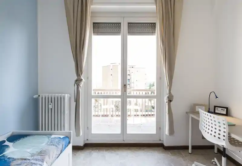 Dormitorio luminoso y minimalista con grandes puertas al balcón que aportan mucha luz natural, cama individual con ropa de cama estampada y un pequeño escritorio — ideal para estudiante o persona sola.