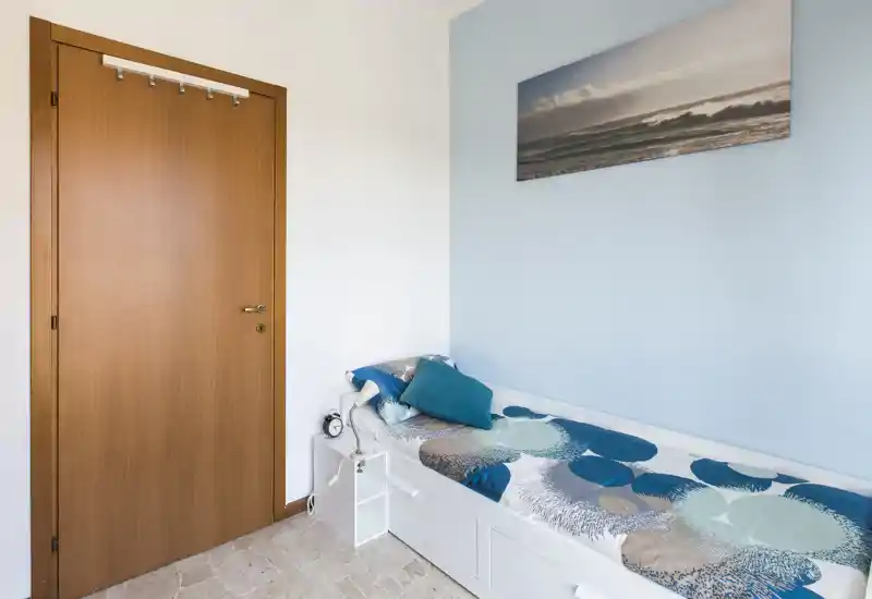 Pequeña habitación individual con cama tipo sofá blanca, cojines en tonos azul y turquesa y cuadro costero sobre pared azul claro. Limpia, ordenada y bien iluminada — ideal para huéspedes o estudiantes.