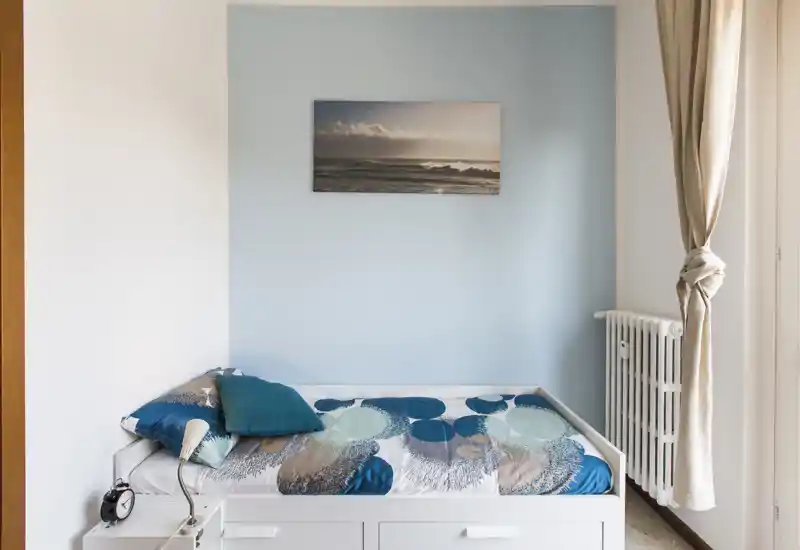 Pequeña habitación luminosa con cama individual contra una pared azul claro, ropa de cama de diseño y cojines, luz natural desde la ventana y estilo minimalista.