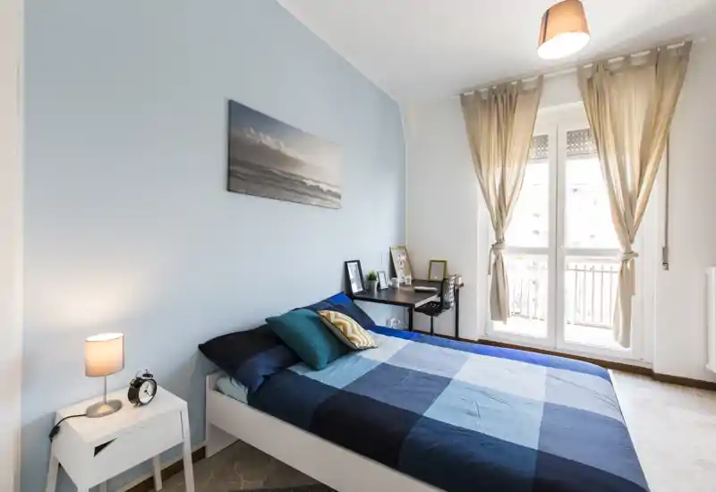 Bright, well-staged bedroom with a double bed dressed in blue linens, a bedside table with lamp and an organized desk by the window letting in abundant natural light — ideal for comfortable stays.