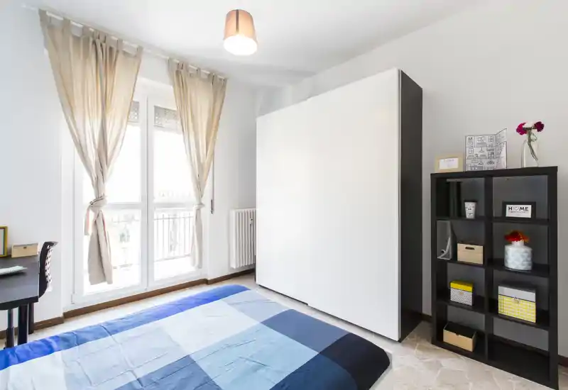 Bright, modern bedroom with a double bed covered in blue geometric bedding, a large white wardrobe, a black cube shelf with decorative storage boxes, and a balcony door with sheer curtains letting in abundant natural light.