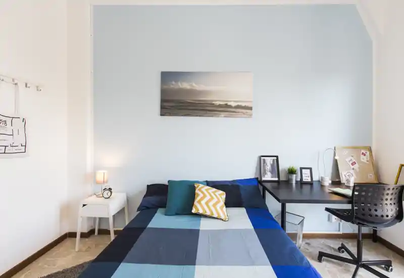 Bright, neatly styled bedroom with a double bed dressed in blue patchwork linens, decorative cushions, a bedside table with lamp, and a workspace desk — ideal for a comfortable stay.