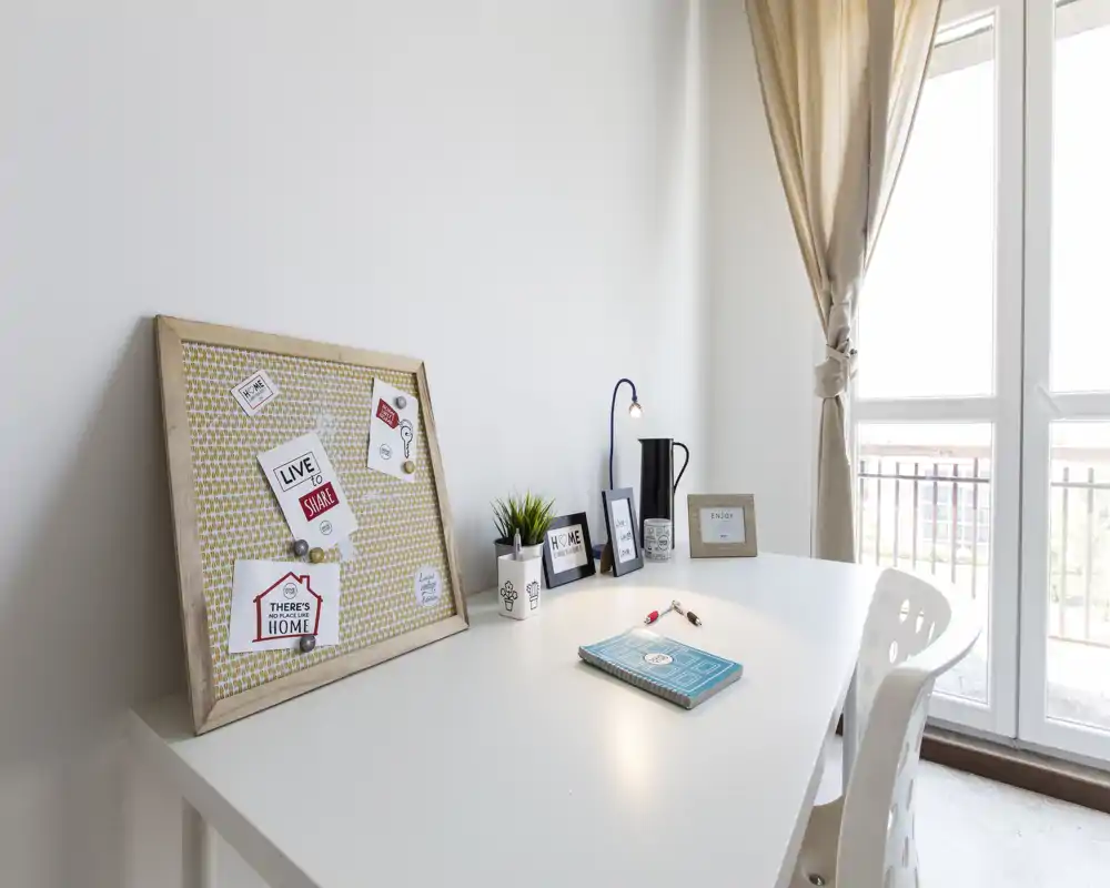 Área de trabajo luminosa y minimalista junto a una gran ventana con un tablón decorativo, una pequeña planta, una lámpara y marcos — un rincón de estudio ordenado y con mucha luz natural.