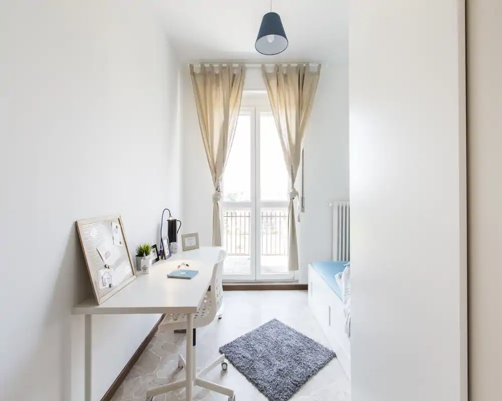 Dormitorio luminoso y minimalista con cama individual, escritorio y puerta-balcon que inunda la estancia de luz natural.