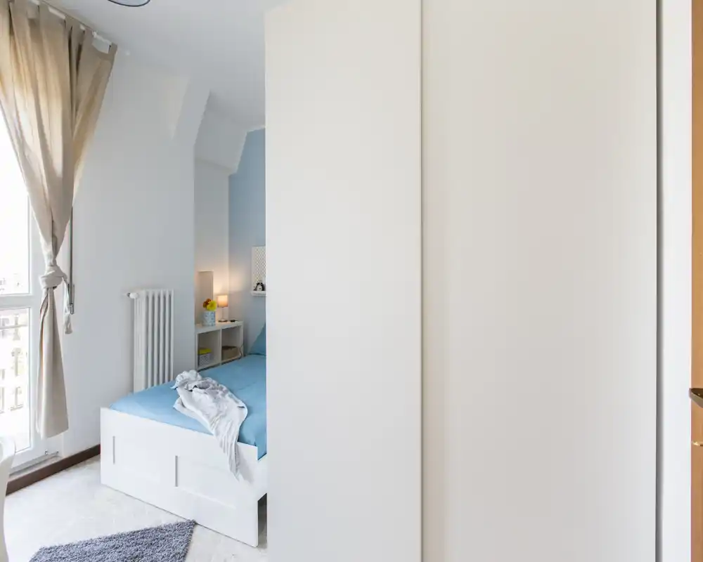 Dormitorio pequeño y luminoso con estructura de cama blanca, ropa de cama azul y mesita con lámpara. La luz natural de la ventana alta y las cortinas neutras crean un ambiente despejado y tranquilo.