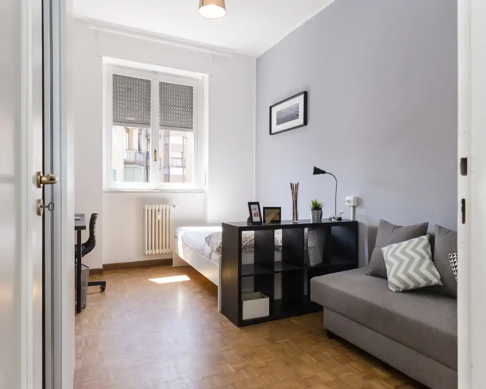 Chambre lumineuse et moderne avec un lit double confortable, un petit canapé et une étagère cubique servant de séparateur. Lumière naturelle provenant d'une grande fenêtre, sol en parquet et décoration minimaliste offrent une atmosphère calme et accueillante, idéale pour la location courte durée.