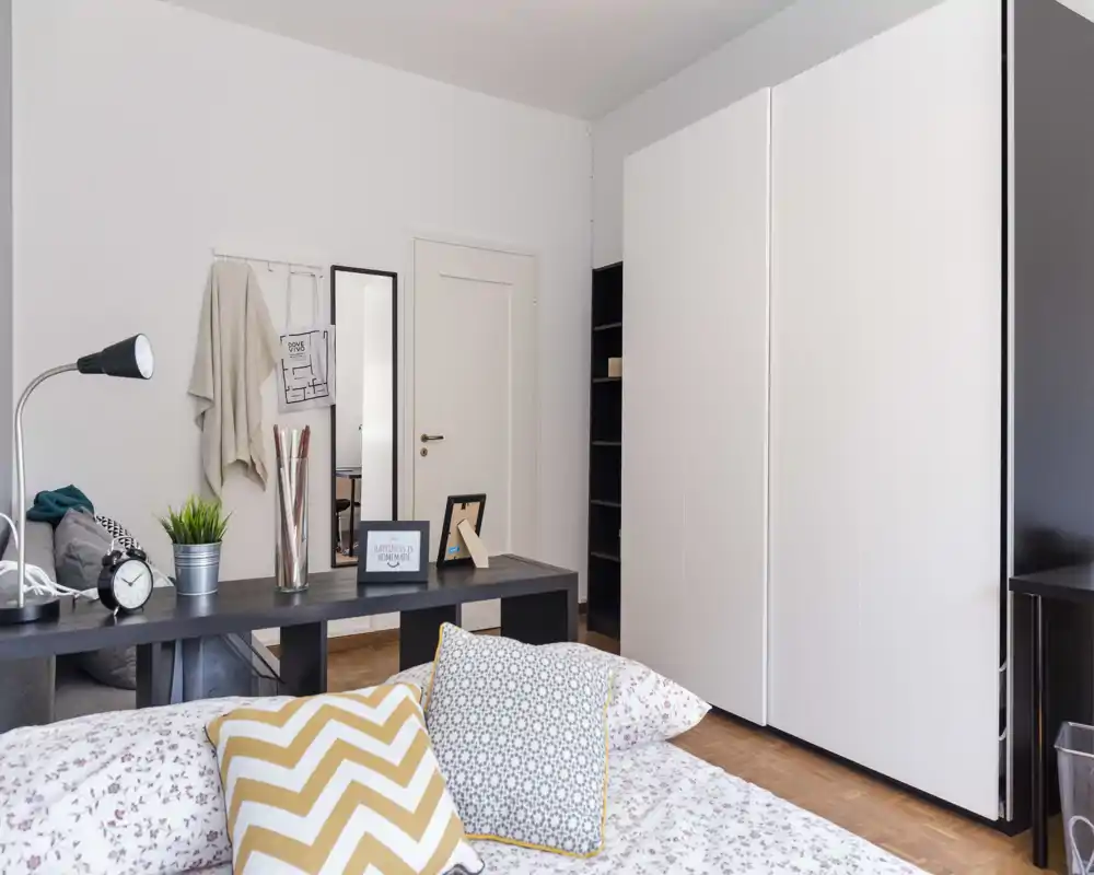 Chambre lumineuse et bien mise en scène avec une literie à motifs et des coussins décoratifs ; grande armoire et bureau derrière le lit pour rangement et travail.