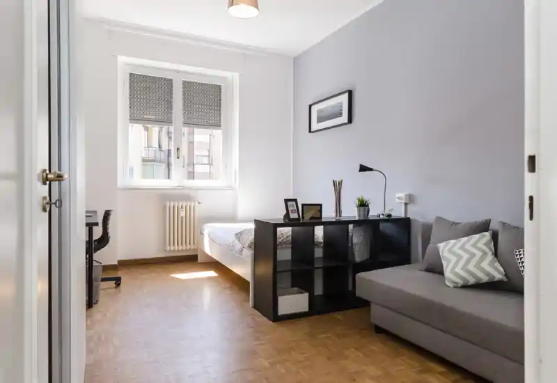 Chambre lumineuse et moderne avec un lit double confortable, un petit canapé et une étagère cubique servant de séparateur. Lumière naturelle provenant d'une grande fenêtre, sol en parquet et décoration minimaliste offrent une atmosphère calme et accueillante, idéale pour la location courte durée.