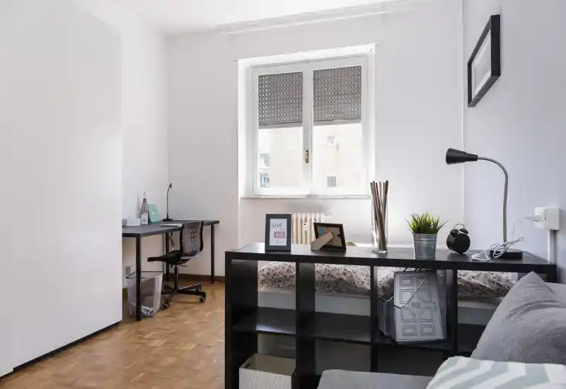 Chambre moderne et lumineuse avec un lit simple derrière une étagère basse, un bureau près de la fenêtre et des éléments décoratifs qui rendent l'espace fonctionnel et chaleureux.