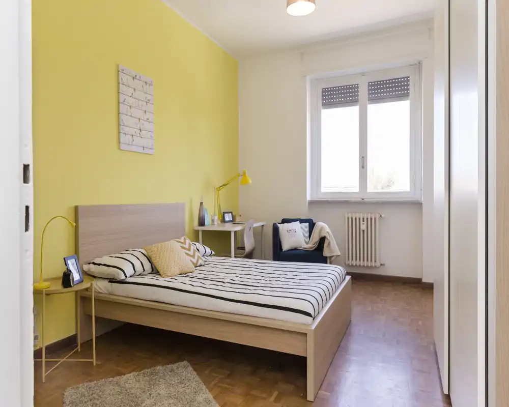 Camera da letto moderna e luminosa con letto in legno chiaro, biancheria a righe e parete d'accento gialla. La stanza dispone di una scrivania, una poltrona e molta luce naturale dalla finestra — perfetta per un soggiorno confortevole.
