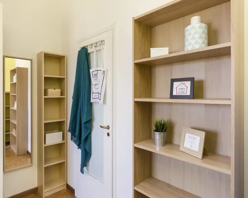 Ingresso piccolo e ordinato con scaffalature in legno chiaro, uno specchio a figura intera e una porta con ganci che reggono una sciarpa verde acqua — ambiente luminoso e minimal.