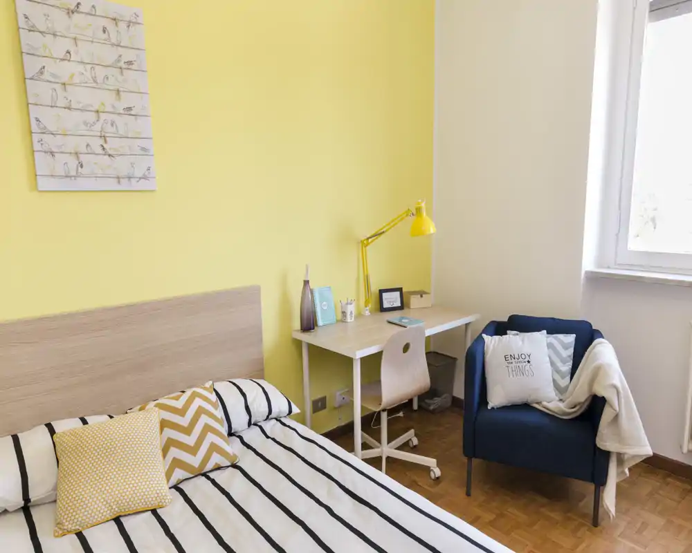 Camera da letto luminosa e ordinata con letto matrimoniale, dettagli gialli coordinati, una scrivania e una poltrona accogliente vicino alla finestra.