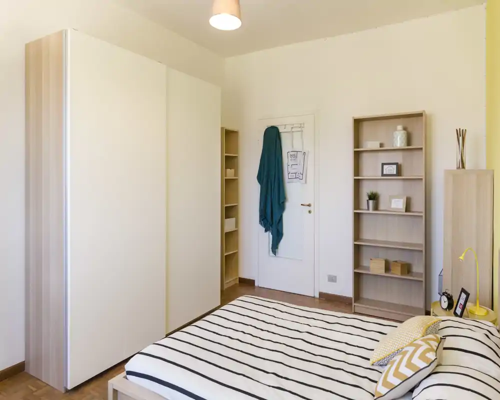 Camera da letto luminosa e ordinata con letto a righe, armadio moderno e scaffalatura aperta. Linee pulite, pavimento in legno caldo e arredi morbidi rendono l'ambiente accogliente per annunci.