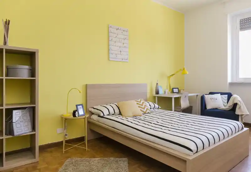 Dormitorio luminoso y acogedor con una cama grande, ropa de cama a rayas y pared amarilla; cuenta con escritorio, sillón y estantería para mayor confort.