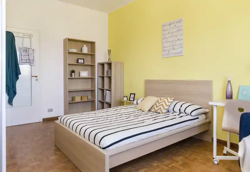 Dormitorio doble luminoso y bien presentado con cama de madera, ropa de cama a rayas y cojines decorativos; estanterías claras y un pequeño escritorio junto a la cama crean un ambiente moderno y acogedor.