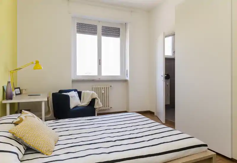 Dormitorio moderno y luminoso con cama doble a rayas, cojines acogedores y un pequeño escritorio junto a la ventana. Decoración sencilla y fresca con un sillón cómodo y luz natural.