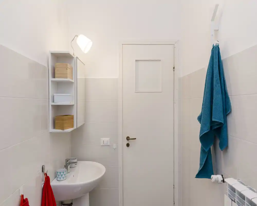 Petite salle de bains propre avec rangement mural, lavabo sur colonne, crochets pour serviettes et carrelage neutre ; lumineuse et bien éclairée.