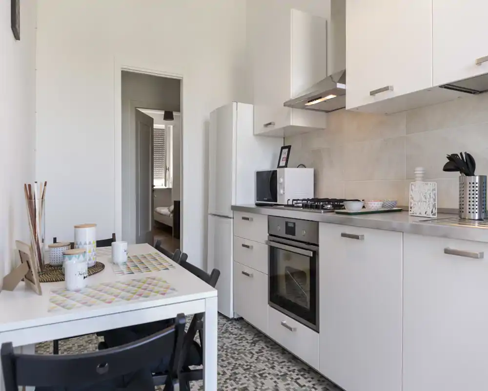 Cuisine compacte et moderne avec meubles blancs, four encastré et plaque gaz, réfrigérateur et petite table à manger; espace lumineux, propre et bien mis en scène, parfait pour une annonce.