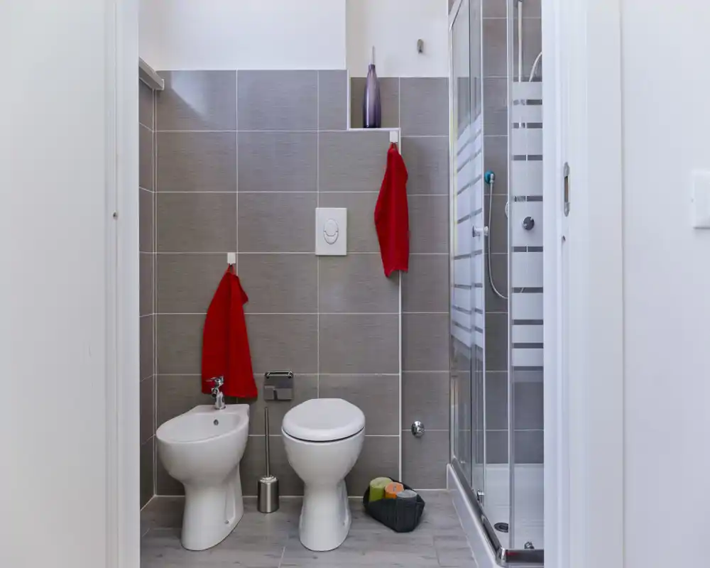 Salle de bain moderne et lumineuse avec plaque de chasse d'eau murale, bidet et WC côte à côte, cabine de douche en verre, carrelage gris et serviettes rouges pour le contraste.