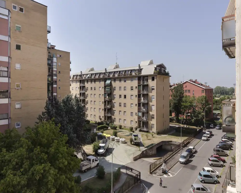 Belle vue extérieure d'un immeuble résidentiel et de la rue depuis un balcon ou une fenêtre en étage, montrant des bâtiments, des voitures stationnées et une petite cour.