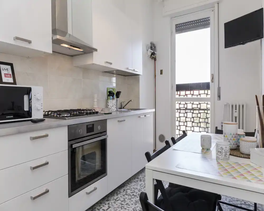 Cuisine moderne et compacte avec placards blancs, four encastré et plaque à gaz, ainsi qu'une petite table à manger près de la porte donnant sur le balcon — idéale pour un appartement en ville.