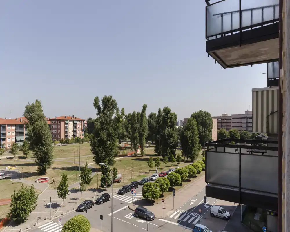 Vue ensoleillée depuis le balcon sur un parc planté d'arbres et une rue résidentielle, avec des balcons d'appartements visibles au premier plan.