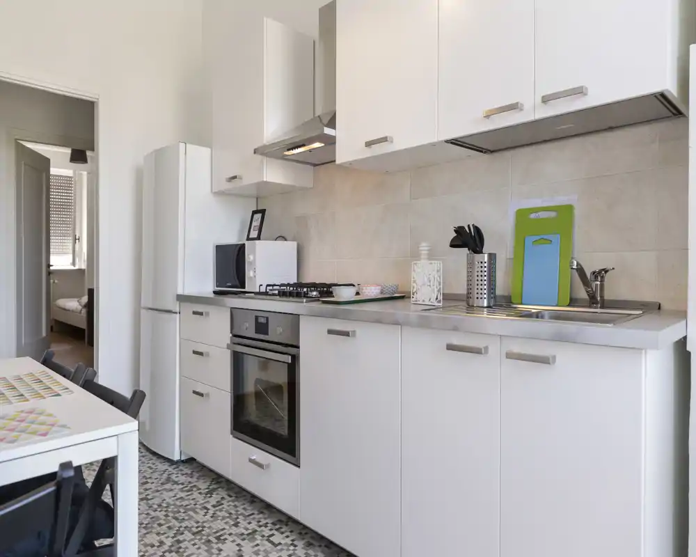 Cuisine moderne et lumineuse avec meubles blancs, four encastré, plaque à gaz, évier et réfrigérateur. Plans de travail propres et accessoires soignés pour un espace de cuisson fonctionnel.