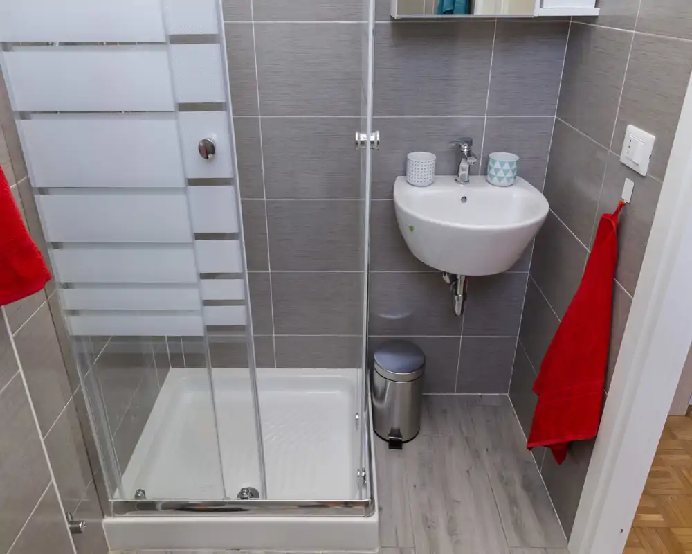 Salle de bains moderne et compacte avec cabine de douche en verre, lavabo mural et serviettes rouges pour une touche de couleur. Carrelage gris et sol effet bois clair.