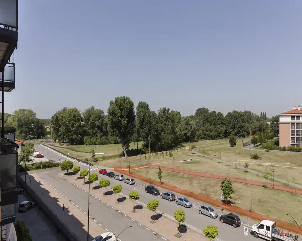 Vue extérieure large depuis un bâtiment : parc bordé d’arbres, sentiers piétons et voitures garées le long de la rue par une journée claire et ensoleillée.