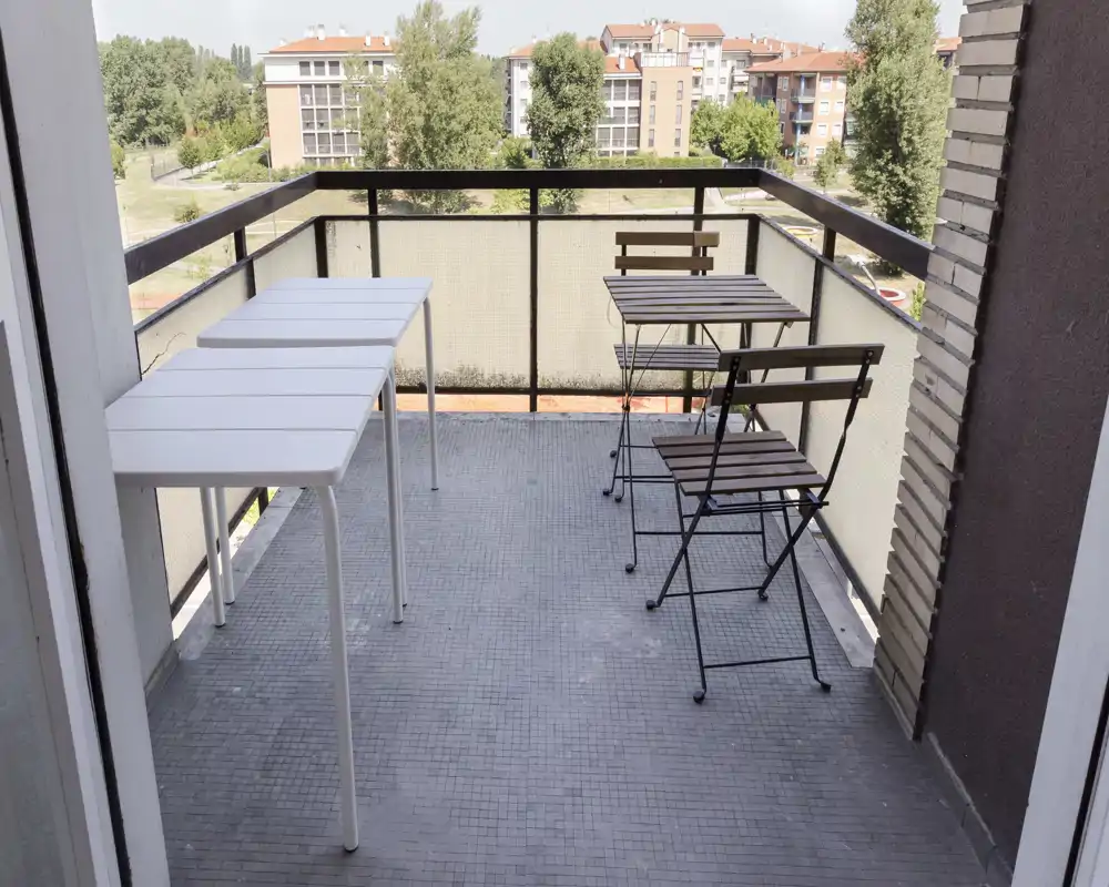 Balcon ensoleillé avec une petite table bistro et deux chaises pliantes, plus deux tables blanches à gauche — idéal pour le café du matin ou un repas léger avec vue sur les bâtiments et la verdure.
