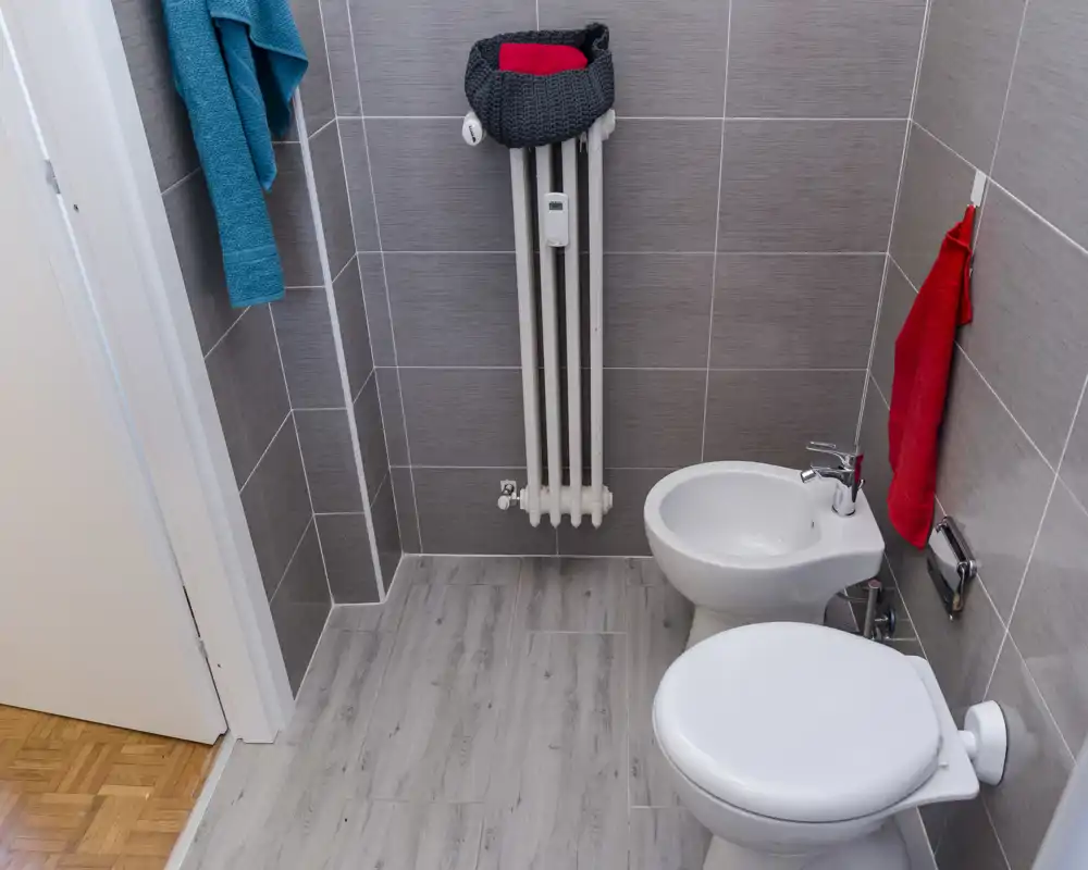 Salle de bain moderne et compacte avec carrelage gris clair, bidet et WC, radiateur mural et serviettes colorées, lumineuse et rangée.