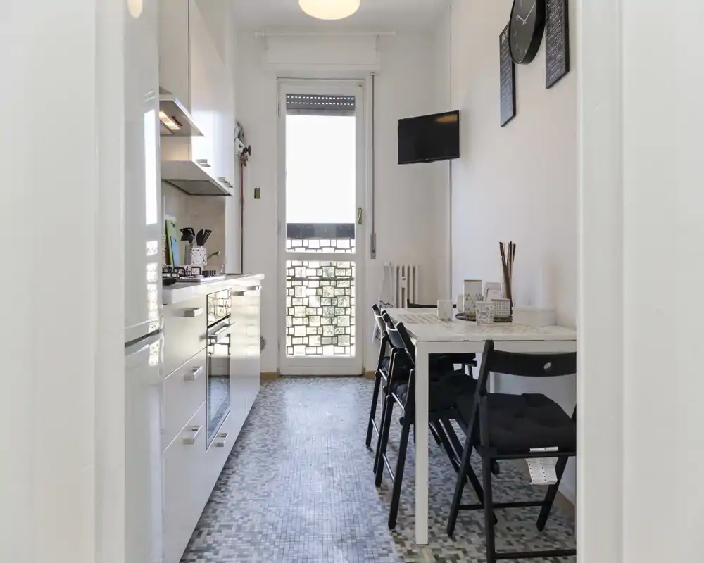Cuisine étroite et lumineuse avec meubles blancs modernes, four encastré et petite table pour quatre près de la porte-fenêtre — idéale pour appartements urbains compacts.