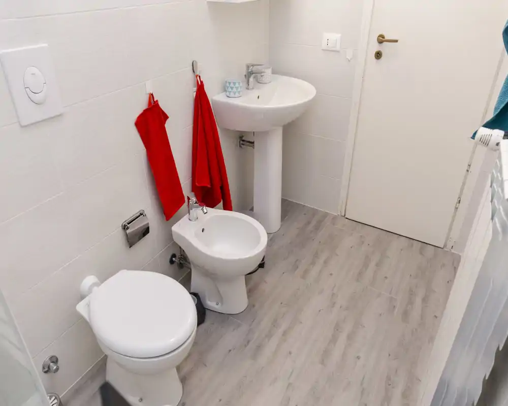 Salle de bains moderne et propre avec WC, bidet et lavabo sur pied, sol effet bois clair et murs carrelés blancs ; serviettes rouges pour une touche de couleur.