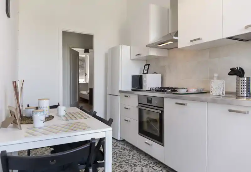 Cucina compatta e moderna con mobili bianchi, forno incassato e piano cottura a gas, frigorifero e piccolo tavolo da pranzo; ambiente luminoso, pulito e ben arredato ideale per annunci.