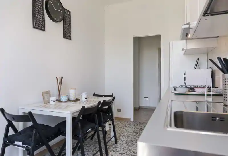 Cucina compatta e luminosa con un piccolo tavolo da pranzo per quattro e lavello in acciaio inox con piano cottura sulla destra. Linee pulite e arredo essenziale ideali per affitti brevi.