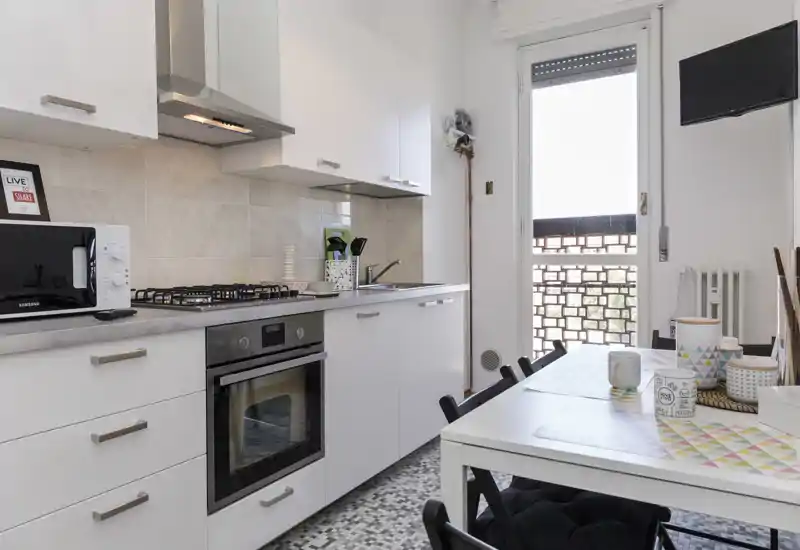 Cucina moderna e compatta con mobili bianchi, forno incassato e piano cottura a gas, più un piccolo tavolo da pranzo vicino alla porta che dà sul balcone — perfetta per un appartamento cittadino.