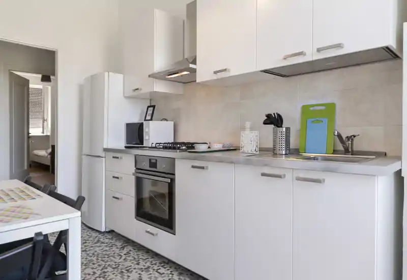 Cucina moderna e luminosa con mobili bianchi, forno integrato, piano cottura a gas, lavabo e frigorifero. Piano di lavoro pulito e accessori curati per uno spazio cucina funzionale.