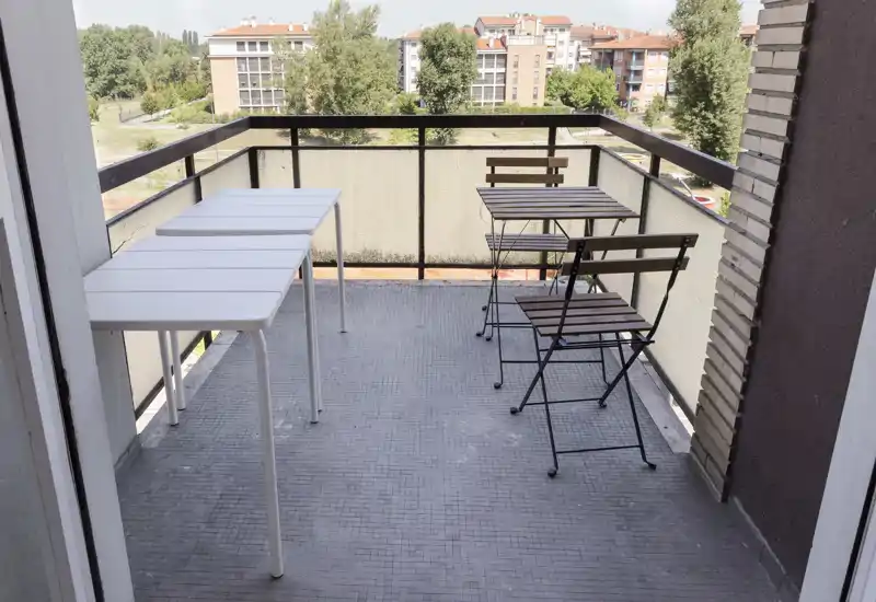 Balcone soleggiato con un piccolo tavolo da bistrò e due sedie pieghevoli, più due tavoli bianchi a sinistra: perfetto per colazione o pasti leggeri con vista su edifici e verde.