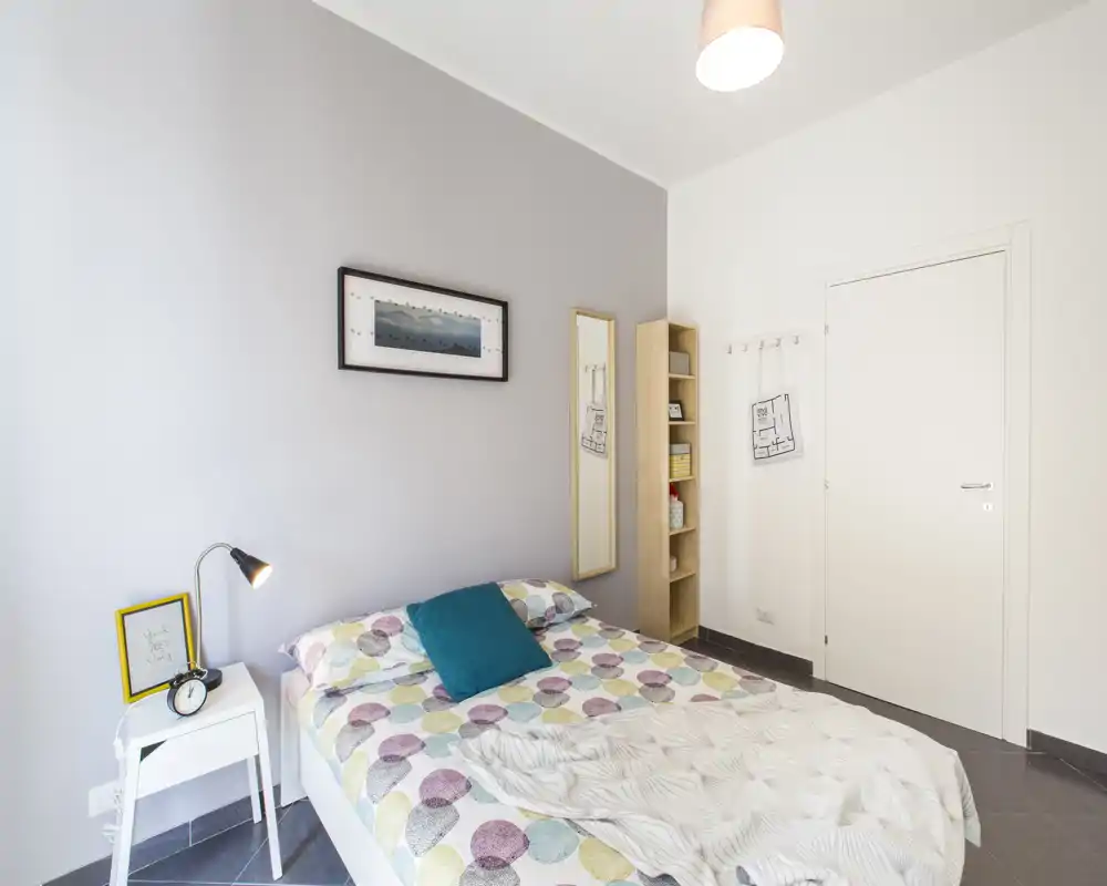 Dormitorio luminoso y moderno con cama doble y ropa de cama de colores, cojín turquesa, mesita con lámpara y espejo vertical; paredes neutras y estantería ordenada completan el conjunto.