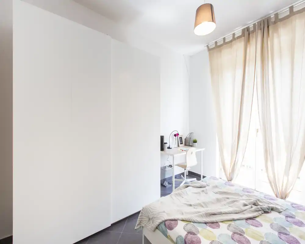 Dormitorio moderno y luminoso con cama doble, textiles claros y amplio armario; un pequeño escritorio junto a la ventana proporciona un práctico espacio de trabajo con luz natural.