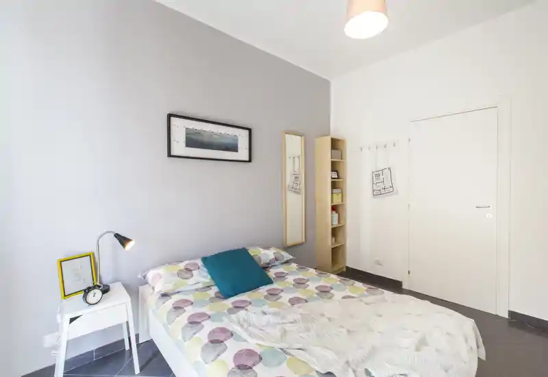 Dormitorio luminoso y moderno con cama doble y ropa de cama de colores, cojín turquesa, mesita con lámpara y espejo vertical; paredes neutras y estantería ordenada completan el conjunto.