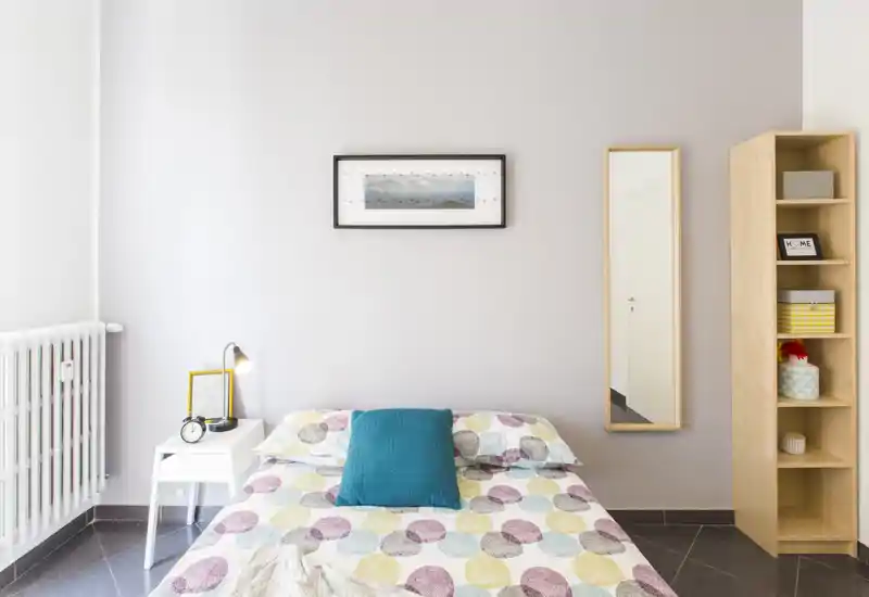 Dormitorio luminoso y minimalista con cama doble, edredón estampado y cojín turquesa; mesita, lámpara y espejo alto completan el ordenado conjunto.