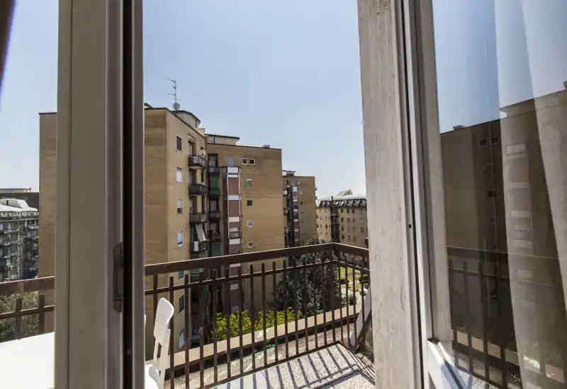 Balcone privato soleggiato con ringhiera in metallo e vista sulla città; accesso tramite porta finestra scorrevole con lieve riflesso interno.