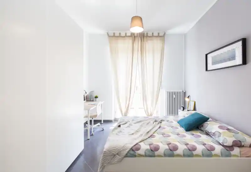 Dormitorio moderno y luminoso con cama doble con ropa de cama colorida, lámpara de noche y una zona de estudio junto a la ventana con abundante luz natural.